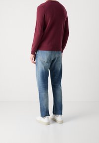 Pull bordeaux à manches longues et col rond, associé à un jean bleu délavé. Des baskets blanches complètent cette tenue décontractée.