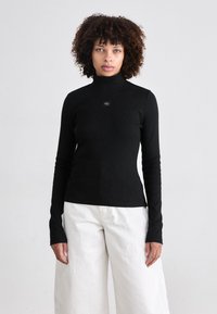 Black long-sleeve turtleneck top with a fitted design, featuring a small logo detail at the chest, paired with white wide-leg trousers.