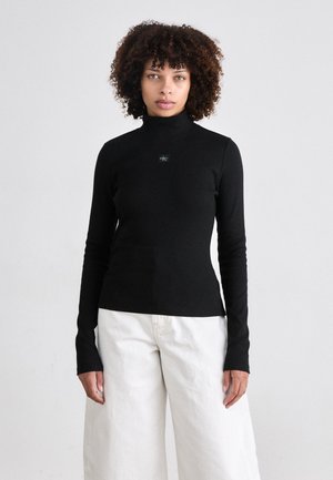 Black long-sleeve turtleneck top with a fitted design, featuring a small logo detail at the chest, paired with white wide-leg trousers.