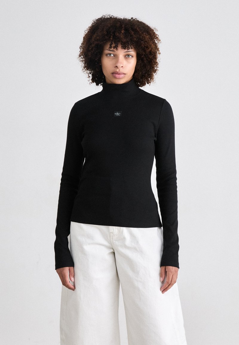 Black long-sleeve turtleneck top with a fitted design, featuring a small logo detail at the chest, paired with white wide-leg trousers.