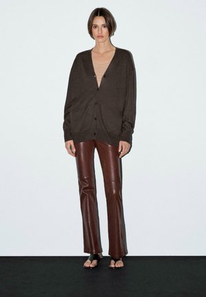 Le mannequin se tient devant un fond uni, portant un cardigan oversized marron, un haut beige et un pantalon en cuir marron, mettant en avant un style minimaliste.