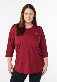 Bordeauxrotes athletisches Langarmshirt mit rundem Ausschnitt, Dreiviertelärmeln und einem silbernen Logoakzent auf der linken Brust. Glatte Stofftextur.