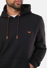 Mustapaita pehmeästä kankaasta, jossa on kengurupocket. Ominaisuuksiin kuuluu oranssit nyörit ja pieni oranssi logo rinnassa.