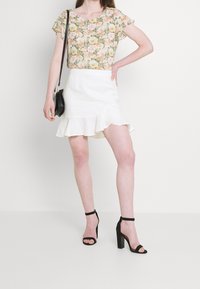 Blusa de manga corta con estampado floral en crema y colores pasteles, combinada con una mini falda blanca con volantes; tacones altos negros con tiras y un bolso.