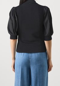 Svart stickad topp med puffade tygärmar, ribbad textur, åtsittande midja och en liten logotypdetalj på baksidan. Kombinerad med ljusblå denimbyxor.