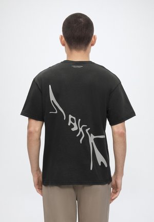 Άνδρας με κοντά σκούρα μαλλιά φοράει μαύρο μπλουζάκι με μεγάλο αφαιρετικό λευκό σχέδιο στην πλάτη, στέκεται μπροστά σε φόντο απλό και φωτεινό.