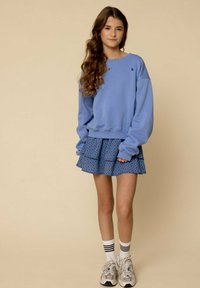 Lichtblauwe sweatshirt met een losvallende pasvorm en geribbelde boorden, in combinatie met een gelaagde blauw patroon rok. Witte sokken met zwarte strepen en grijze sneakers.