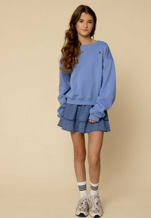 Lichtblauwe sweatshirt met een losvallende pasvorm en geribbelde boorden, in combinatie met een gelaagde blauw patroon rok. Witte sokken met zwarte strepen en grijze sneakers.