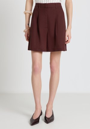 Femme portant une jupe plissée marron foncé jusqu'au genou, un haut rayé et des chaussures pointues marron foncé, debout devant un fond uni.