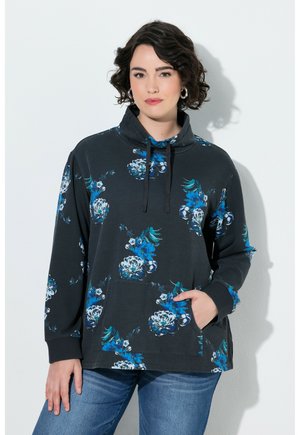 Kvinde med kort krøllet hår iført en sort sweatshirt med blå og hvide blomsterprint og blå jeans, stående med den ene hånd i lommen.