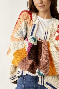 Γυναίκα με πολύχρωμο χοντρό πλεκτό πουλόβερ που κρατάει μια πλεκτή κουβέρτα πατσγουόρκ με διάφορα χρώματα.