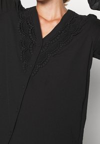 Blouse noire avec une bordure en dentelle décorative le long du décolleté en V, dotée d'une fermeture à bouton et de manches courtes. Texture lisse dans l'ensemble.