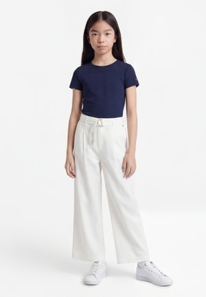 Niña de pie con una camiseta de manga corta azul marino, pantalones blancos de pierna ancha de cintura alta con cinturón y zapatillas blancas sobre un fondo blanco.