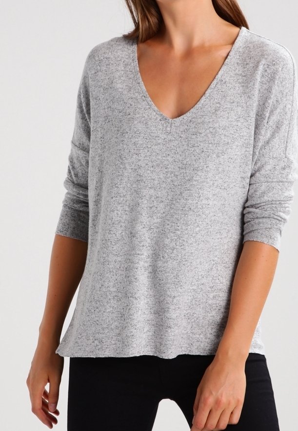 Femme portant un pull gris clair à col en V et manches longues avec une texture douce, et un pantalon noir sur un fond uni.