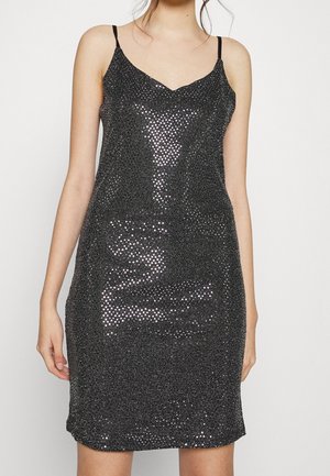 Zwart pailletten slipjurk met dunne bandjes gedragen door een persoon, valt boven de knieën tegen een effen lichte achtergrond.