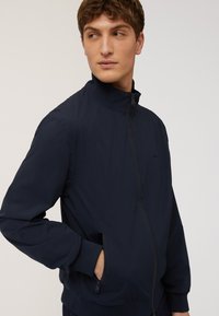 Giacca leggera blu navy con colletto alto, zip frontale e tasche laterali. Texture morbida con polsini e orlo a coste. Branding discreto.
