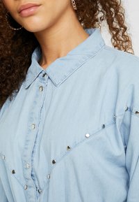 Light blue denim shirt featuring a classic collar, metallic snap buttons, and decorative studs along the front and shoulders.