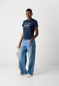 Marineblå bomulls-T-skjorte med hvit merkevare, kombinert med lyseblå vide denimjeans. Hvite sneakers fullfører det avslappede utseendet.