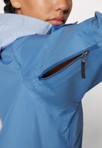 Chaqueta azul con una tela suave y texturizada, que presenta un bolsillo con cremallera lateral y un forro interior en contraste. El diseño enfatiza la funcionalidad y el ajuste.