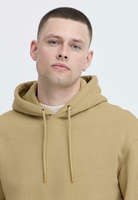 Tan hoodie med snor, tekstureret stof og en kengurulomme. Båret over en lysefarvet skjorte. Enkel, afslappet design.