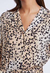Chemise à imprimé léopard beige et noir, en tissu léger, avec col, manches courtes et fermeture à boutons sur le devant. Détail de taille élastique.