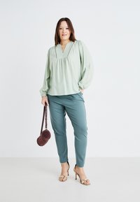 Light green blouse with a v-neck and gathered detail, paired with teal pants. Accessories include a round, dark purple bag and gold heeled sandals.