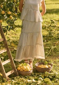 Femme en robe à carreaux debout près des citronniers avec deux paniers remplis de citrons et une échelle en bois sur un sol herbeux.