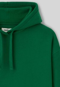 Sweat à capuche vert en tissu doux. Il dispose d'un cordon de serrage au col et d'une surface texturée, sans logos ni motifs visibles.