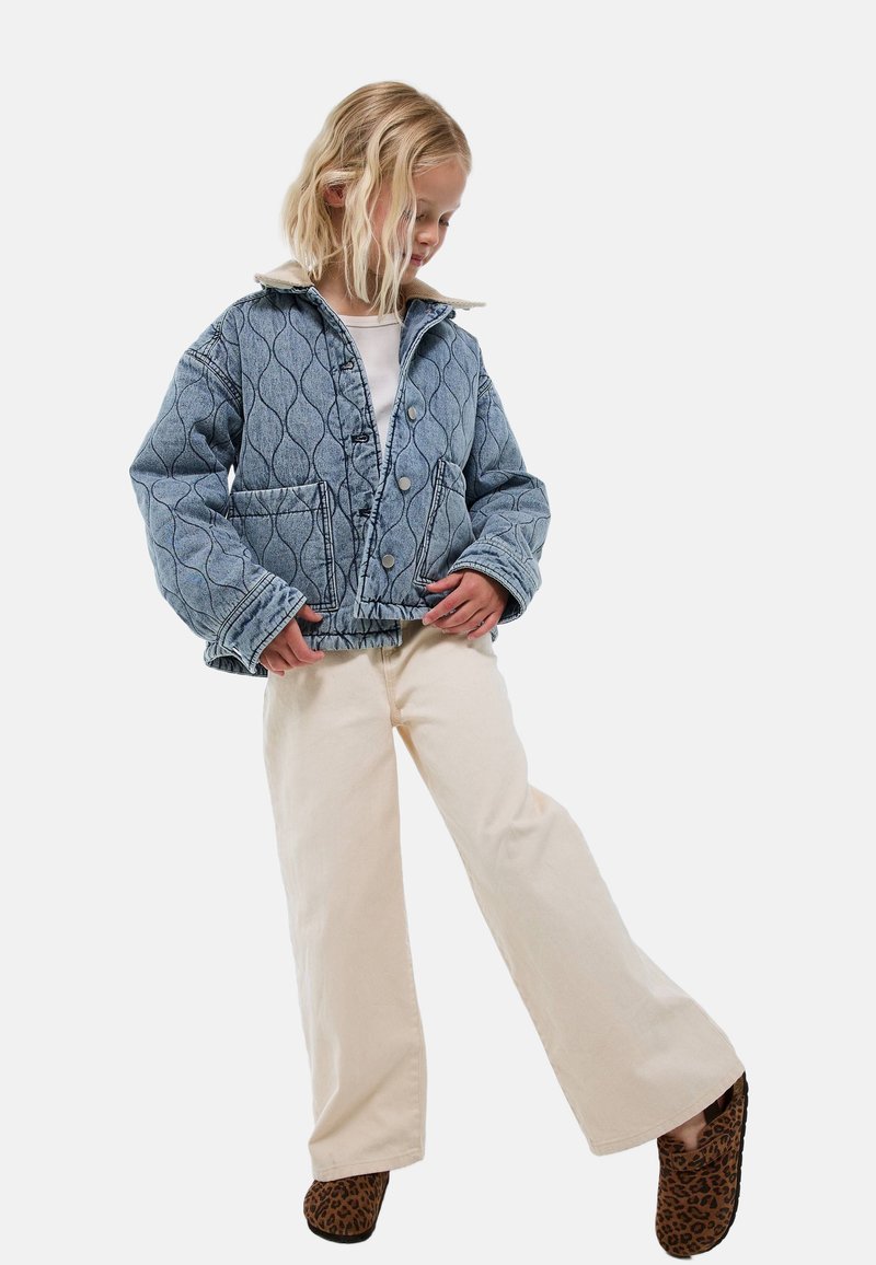 Veste en denim avec un motif matelassé, couleur bleu clair, fermetures à boutons et col. Associée à un pantalon large crème et des chaussons à imprimé léopard.