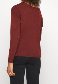 Roter Strickpullover mit lockerer Passform, strukturierten Ärmeln und weitem Ausschnitt. Kombiniert mit schwarzen Jeans, die einen lässigen Stil zeigen.