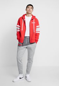 Felpa rossa con zip e coulisse bianche, accenti grigi, abbinata a pantaloni della tuta grigi con il logo rosso di Nike, indossata con sneaker bianche.