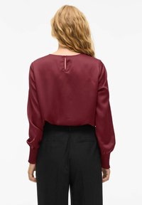 Camicia bordeaux con maniche lunghe, trama di satin morbido, dettaglio con bottoni sul retro, abbinata a pantaloni neri a vita alta.