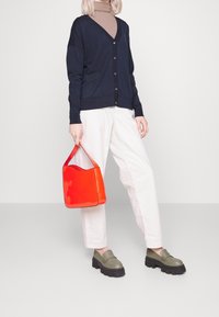 Marinblå cardigan med knäppning, krämfärgade byxor, gröna slipskor och en glänsande röd väska, framför en enfärgad bakgrund.