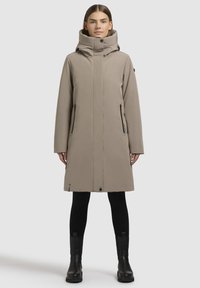 Beige jas met een hoge kraag, lange mouwen, twee zijzakken met rits en een rechte snit. Zwarte accenten nabij de ritsen en de zoom.