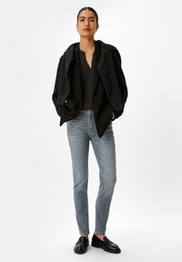 Schwarzer Blazer mit strukturiertem Kragen, kombiniert mit einem dunklen Oberteil, hellgrauen Jeans und glänzenden schwarzen Slippern. Schlichtes Design mit subtilen Details.
