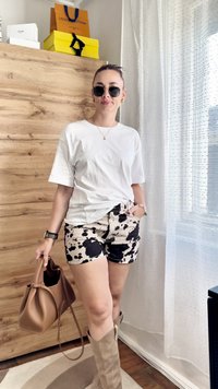 Valkoinen puuvillapaita, musta ja kermaisarja lehmäkuvioiset shortsit, ruskea käsilaukku, beige polvisukat. Luonnollinen puutausta ja läpikuultavat verhot.