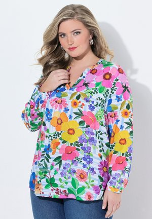 Kvinde med langt blondt hår iført en farverig blomstret bluse med pink, gule og lilla blomster samt blå jeans mod en hvid baggrund.