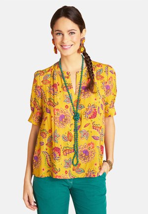 Smiling woman with dark braid wearing yellow floral blouse, teal pants, green beaded necklace, flower-shaped earrings, and colorful bracelet.