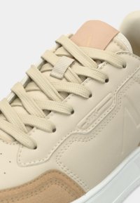 Sneaker beige avec une tige en cuir texturé, des accents en daim, des lacets ton sur ton et une semelle en caoutchouc blanche. Détail de marque subtil présent.