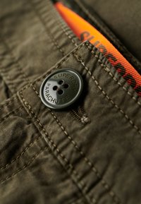Groene stof met zichtbare textuur, voorzien van een opvallende donkere knoop met de opdruk "SUPERDRY PARACHUTE" en een oranje label dat naar buiten steekt.