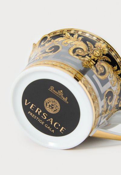 Κύπελλο Versace Prestige Gala, με περίτεχνα χρυσά και μαύρα λουλουδένια σχέδια, διαφανές γυάλινο σώμα και λευκή βάση με λογότυπο της μάρκας.