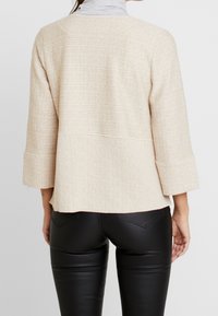 Bežový textured svetr s 3/4 rukávy a vykrojeným lemem, nošený přes černé přiléhavé kalhoty. Materiál vypadá měkký a lehký.