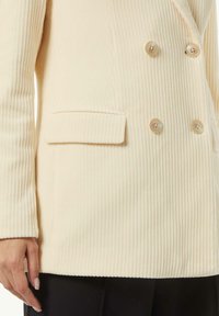 Cremfarbener Cordblazer mit doppelter Knopfleiste, zwei Knöpfen und einer einzelnen Brusttasche. Die Textur ist gerippt.