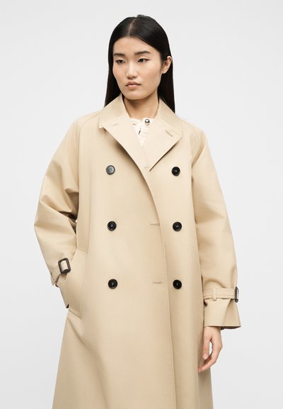 Γυναίκα με ίσια σκούρα μαλλιά που φοράει ένα μπεζ διπλόμπουτο γκρι πανωφόρι με μαύρα κουμπιά και μανσέτες με ζώνη, στέκεται μπροστά σε απλό φόντο.