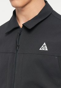 Casaco preto com fecho à frente, gola pontiaguda e logótipo triangular branco. Apresenta um tecido texturizado e detalhes de costura em contraste.