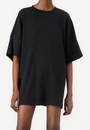 Personne portant une robe noire à manches courtes et ample avec un col rond, debout devant un fond blanc uni.