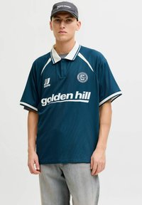 Homme portant un polo de sport bleu marine avec le texte "golden hill" et une casquette noire, debout devant un fond clair uni.