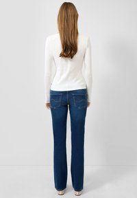 Frau mit langen Haaren, die ein weißes Langarmshirt, blaue Jeans mit geradem Bein und weiße offene Schuhe trägt, vor einem einfarbigen Hintergrund stehend.