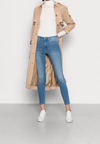 Mujer vestida con un abrigo beige, suéter blanco de cuello alto, jeans ajustados azul claro y zapatillas blancas, de pie con una mano en el bolsillo.