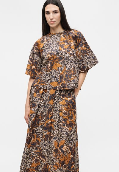 Γυναίκα με χαλαρό, ταιριαστό τοπ και φούστα με περίπλοκο πορτοκαλί και ναυτικό μπλε λουλουδάτο σχέδιο, στέκεται μπροστά σε απλό φόντο.